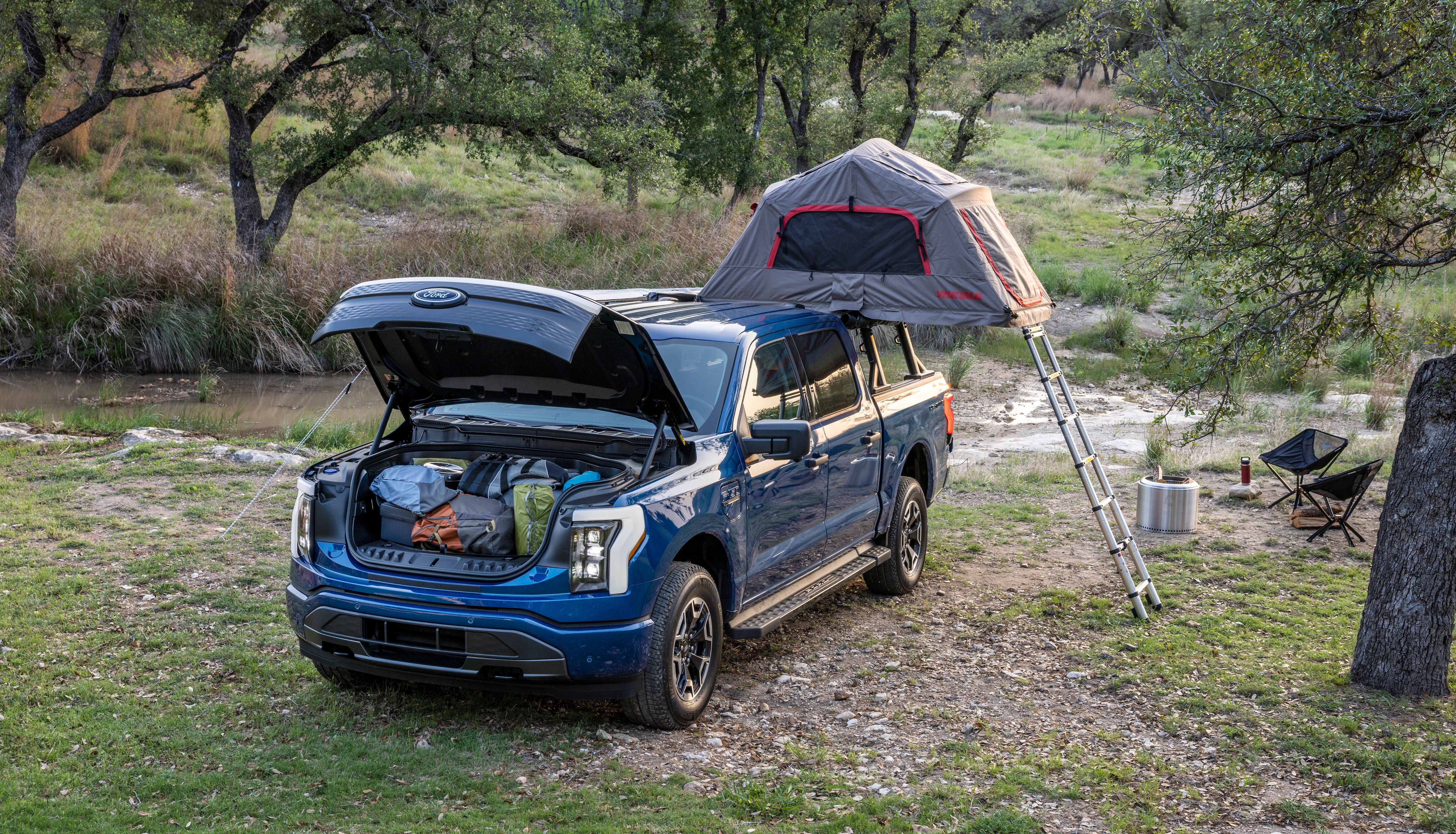 The frunk of a Ford electric truck shown packed with camping supplies as the truck is parked at a campsite with a tent on top of it